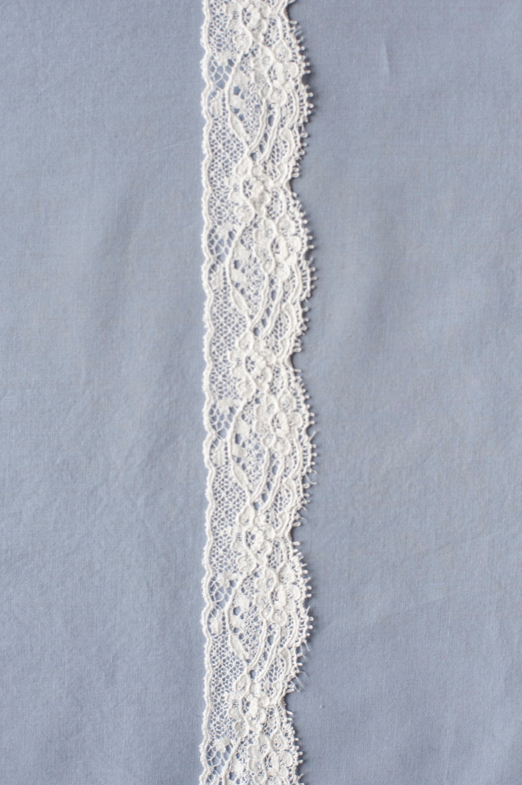 Off-white cotton lace with ornate floral scroll embroidery and openwork pattern, featuring curved edges and detailed texture.