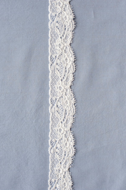 Off-white cotton lace with ornate floral scroll embroidery and openwork pattern, featuring curved edges and detailed texture.