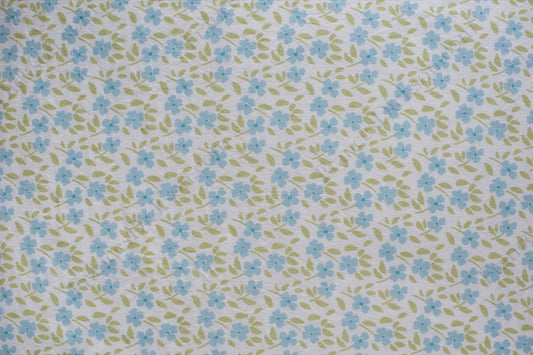 Organic cotton voile fabric with small blue flowers and green leaves scattered across a white background in a ditsy floral print.