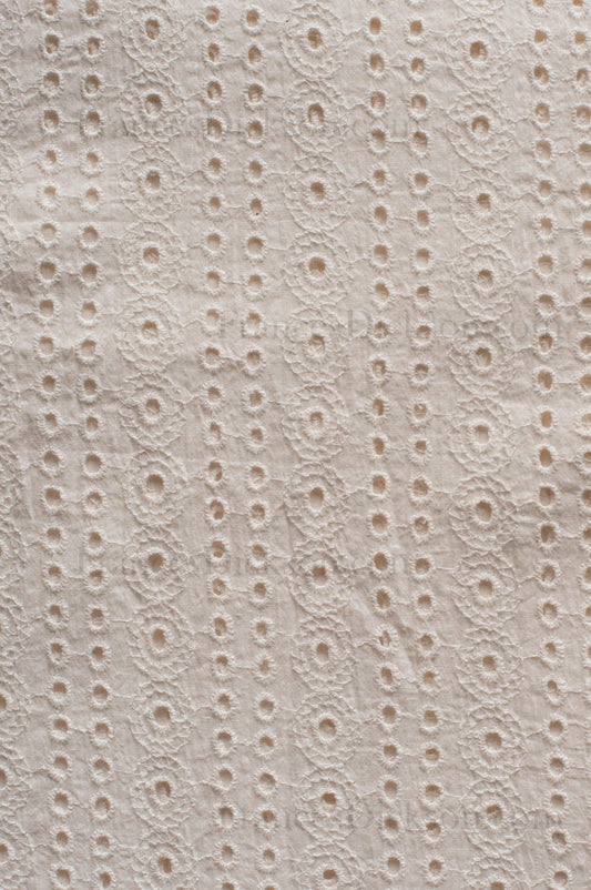 Ivory cotton Schiffli fabric with vertical eyelet embroidery pattern and floral detailing, showing breathable cutwork texture.
